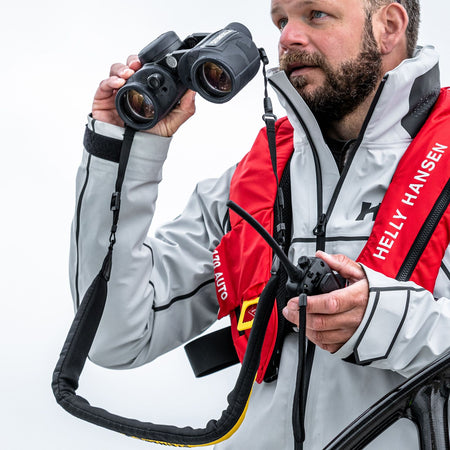 Steiner Schwimmtragegurt für Navigator – Segler mit Helly-Hansen-Schwimmweste blickt durch STEINER Marine-Fernglas an Bord eines Segelboots