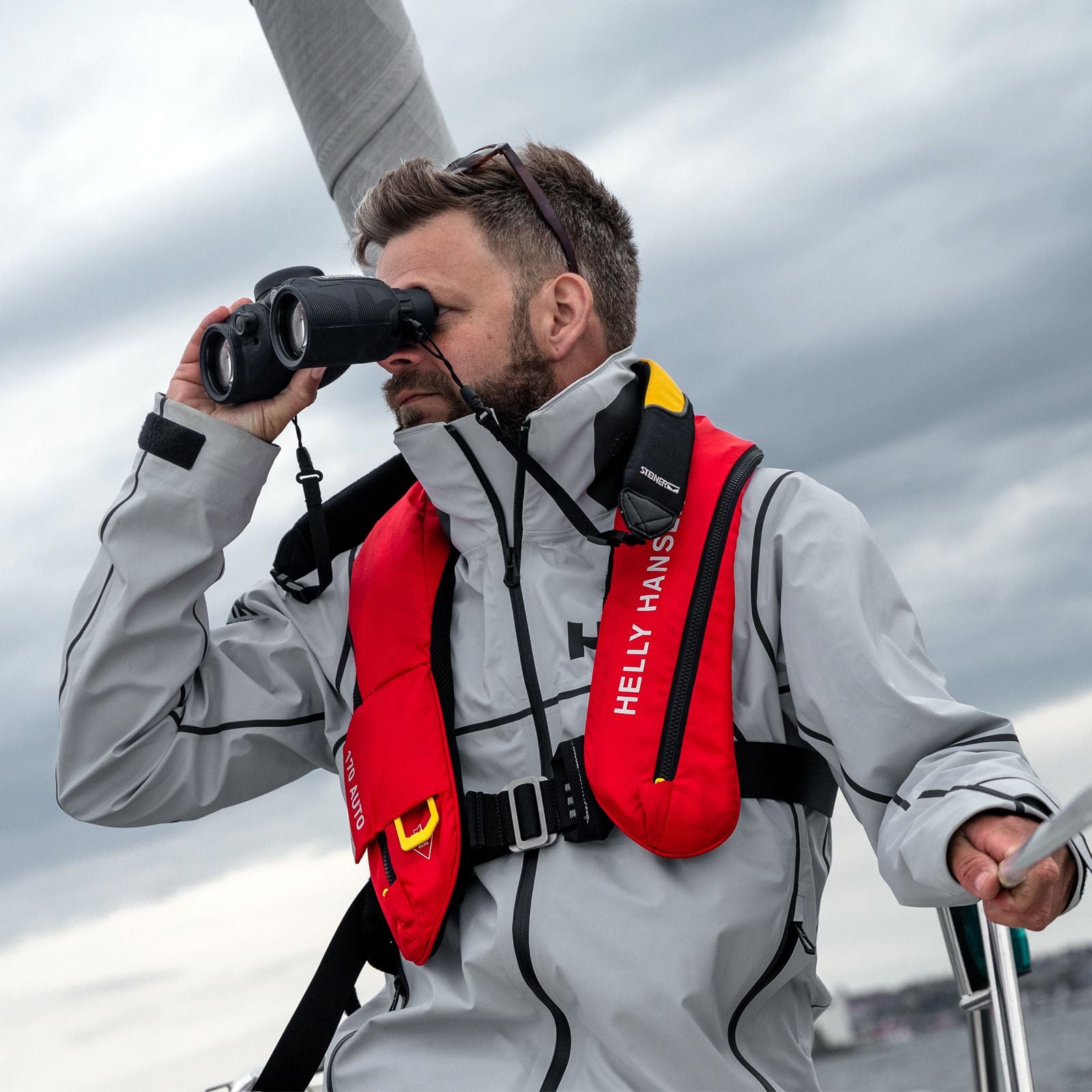 Steiner Schwimmtragegurt für Navigator – Segler in Helly-Hansen-Ausrüstung hält STEINER Marine-Fernglas mit montiertem Schwimmgurt, Funk-Gerät in der anderen Hand