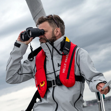 Steiner Schwimmtragegurt für Navigator – Segler in Helly-Hansen-Ausrüstung hält STEINER Marine-Fernglas mit montiertem Schwimmgurt, Funk-Gerät in der anderen Hand