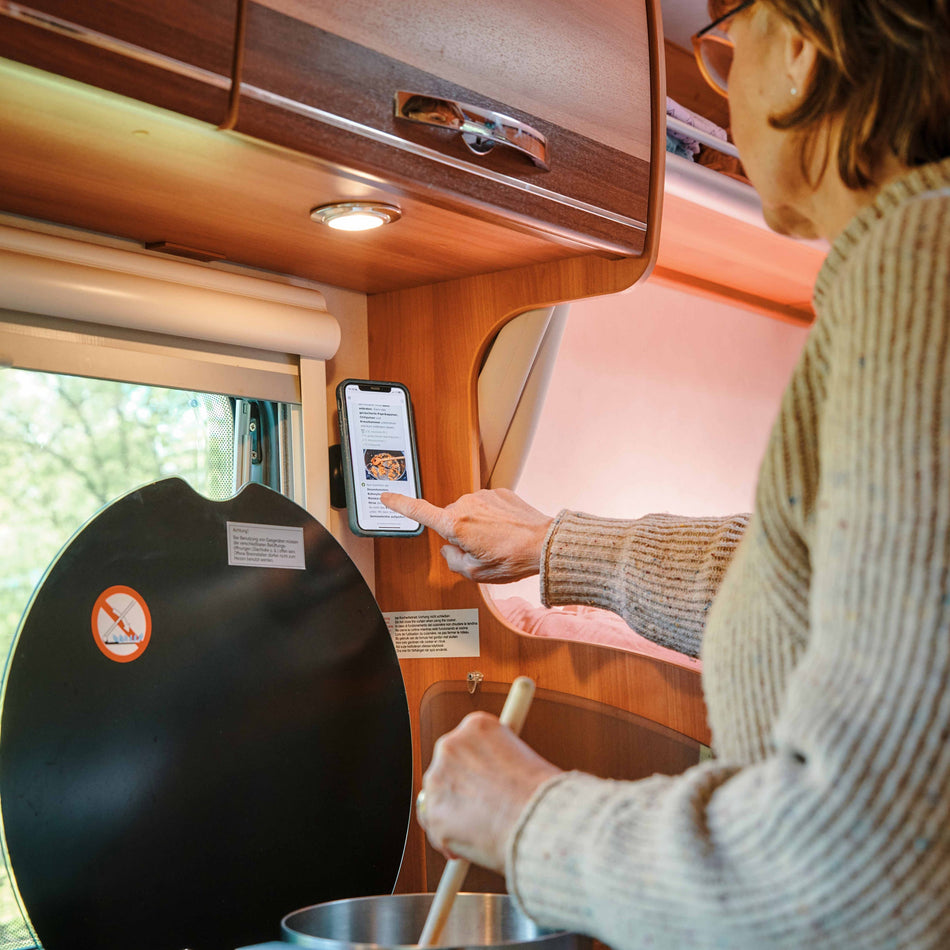 FIDLOCK VACUUM wall base im Einsatz – Frau bedient Smartphone in FIDLOCK-Halterung an Wohnmobilwand beim Kochen
