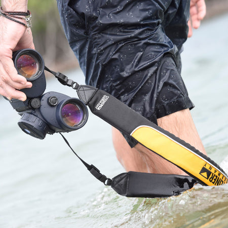Steiner Schwimmtragegurt für Commander 7x50 – Hand hält STEINER Commander-Fernglas mit montiertem gelbem Schwimmgurt über Wasser, nasses Gelände im Hintergrund