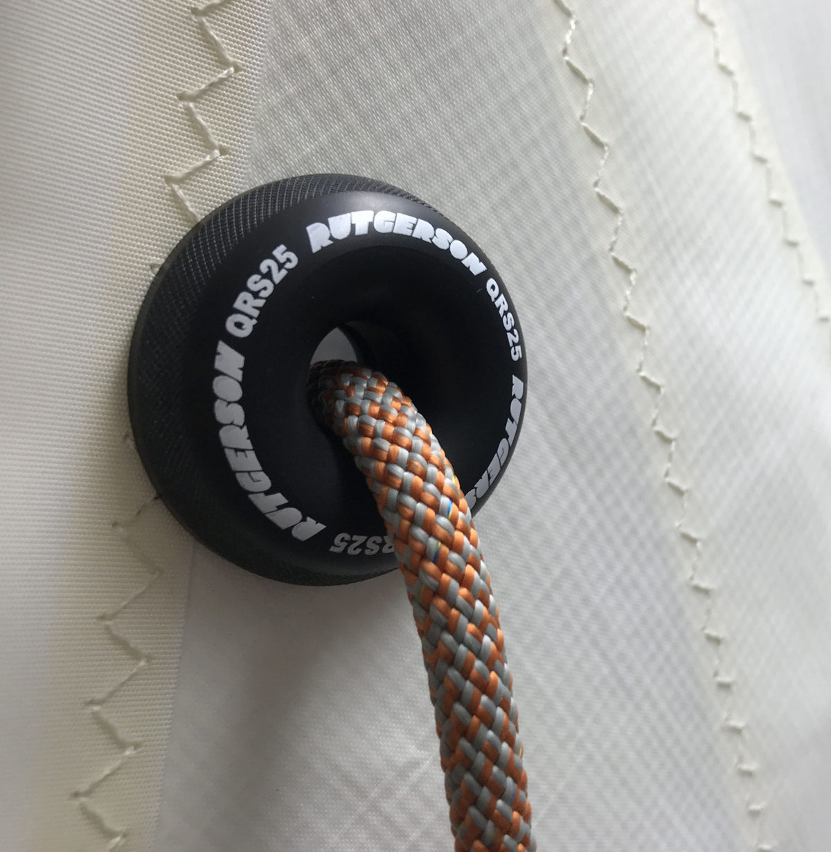 RUTGERSON Quick Reef Ring QRS25 am weißen Segeltuch montiert, mit weißem Markenaufdruck und einer grau-orange geflochtenen Reffleine durch die zentrale Öffnung.